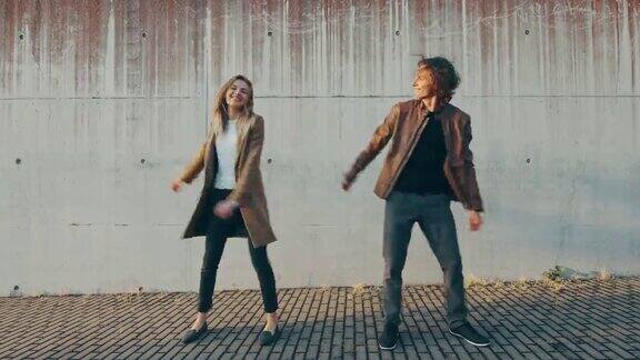 在城市混凝土墙旁边的街道上欢快的女孩和快乐的长发青年正在积极地跳舞他们穿着棕色的皮夹克和外套阳光明媚的一天