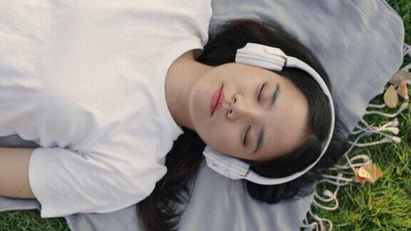 亚洲女性戴着耳机听音乐而躺在地板上的草地在一个公园的户外美丽的日落