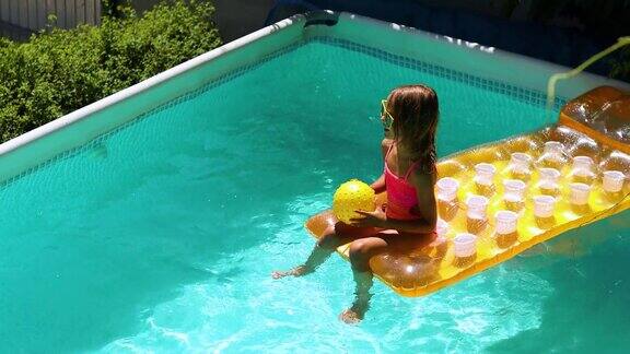 小女孩坐在黄色充气床垫上玩球在游泳池里玩得很开心