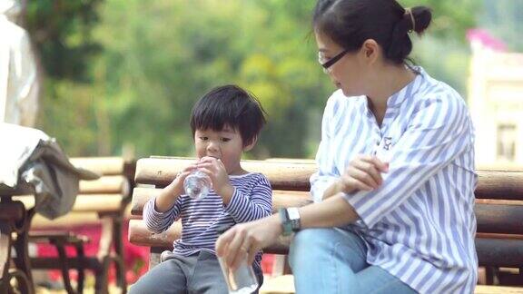 亚洲母亲和她的儿子坐在公园的长凳上喝水