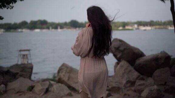 后视镜资深白人妇女长棕色头发站在湖岸欣赏美丽的自然日落体贴、苗条、优雅的女士在有风的夜晚享受户外的休闲