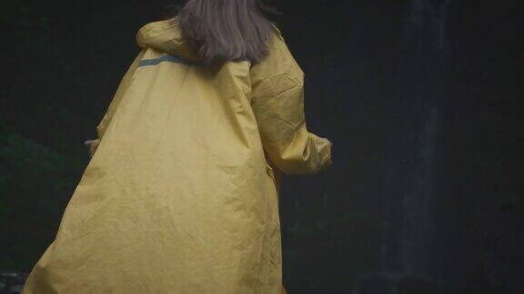 身穿黄色雨衣的少女徒步旅行者奔向瀑布举起双手享受大自然和生活