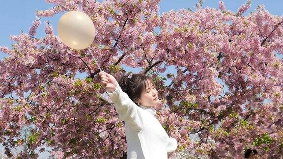美丽的年轻女子双臂张开手拿一个气球站在粉红色的樱花树旁享受风吹过她的脸和头发闭上眼睛4k视频慢镜头