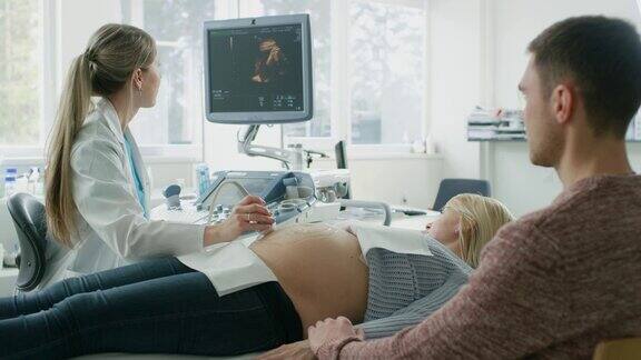 在医院里产科医生使用换能器对孕妇的腹部进行超声超声波扫描丈夫支持妻子并握着她的手