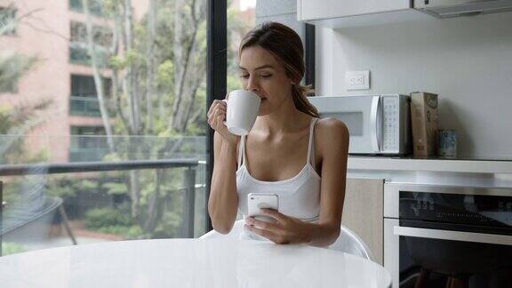 美丽的拉丁美洲女人放松在家查看她的社交媒体同时喝咖啡