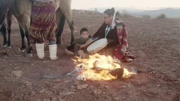 视频剪辑的两个印第安姐妹和他们的小弟弟传统着装纳瓦霍毯子周围他们保持温暖的篝火他们的马和标志性的纪念碑和日落在背景