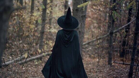 在秋天的森林里一个无法辨认的女人像黑女巫一样在树木之间行走穿着长裙披着斗篷戴着仙女帽的女孩万圣节概念cosplay装扮