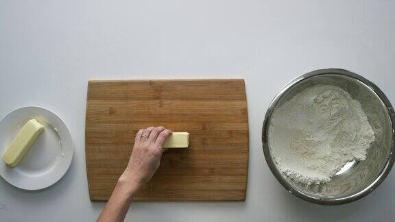 一名年轻女子的手从盘子里移动一根黄油到白色桌子上的木砧板上桌子上还有一个搅拌碗里面有面粉