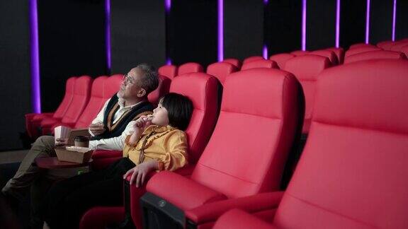 亚裔华裔爷爷和孙女坐在空荡荡的电影院的红色座位上一边吃一边看电影