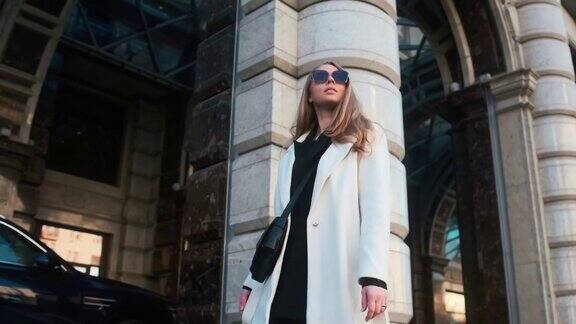 一个戴着眼镜穿着白色外套的女孩走过城市的商业区在豪华轿车和公寓的背景下