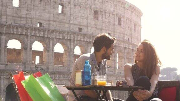 快乐的年轻夫妇游客聊天放松有乐趣坐在酒吧餐厅在罗马斗牛场前在日落与咖啡果汁和购物袋