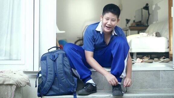 快乐的亚洲学校男孩在运动制服领带蝴蝶结为他的鞋带前在家里上学