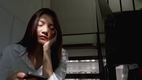 沮丧的年轻美丽的亚洲女人独自坐在楼梯上用智能手机上网有压力的孤独少女盯着手机带着负面情绪然后闭上眼睛