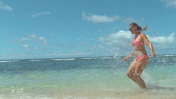 慢镜头:身着粉色比基尼的快乐女孩在海滩边奔跑溅起水花