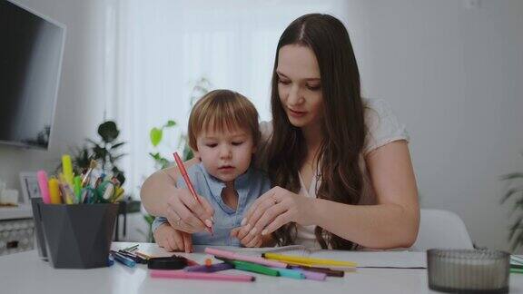 一个年轻的母亲坐在客厅的桌旁教她的儿子拿铅笔教他画画