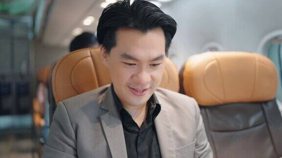 快乐的亚洲英俊商人穿着西装用笔记本电脑坐在飞机里