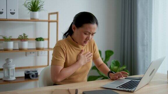 一名亚洲女性坐在家里的办公室里在笔记本电脑前工作患有胸痛