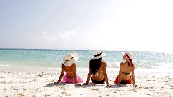 后视图三个女孩在海滩上穿着比基尼和草帽享受日光浴谈话女性游客在暑假
