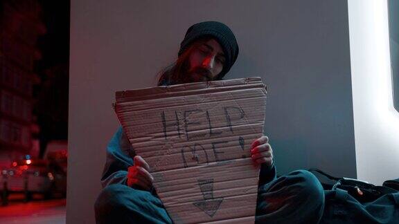 一个留着胡子和长发的无家可归的乞丐坐在街上寻求帮助的慢动作拿着“帮我”的牌子无家可归4k颜色分级