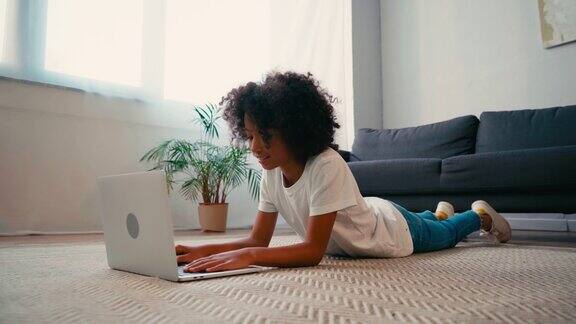 一个非裔美国女孩躺在家里的地毯上在笔记本电脑上打字
