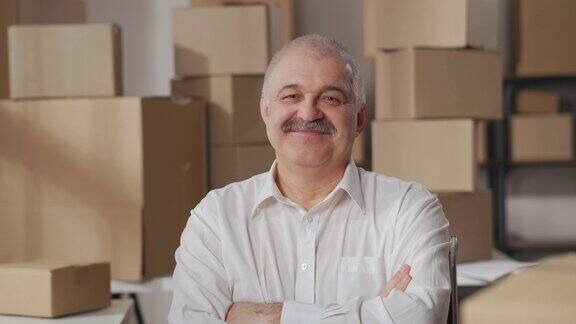 小企业主的肖像一个成熟的男人与胡子在白衬衫交叉他的双臂看着和微笑用纸板箱装货物的仓库