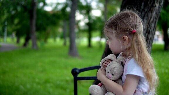 在一个阴天里一个小女孩带着一只玩具兔子坐在公园的长椅上
