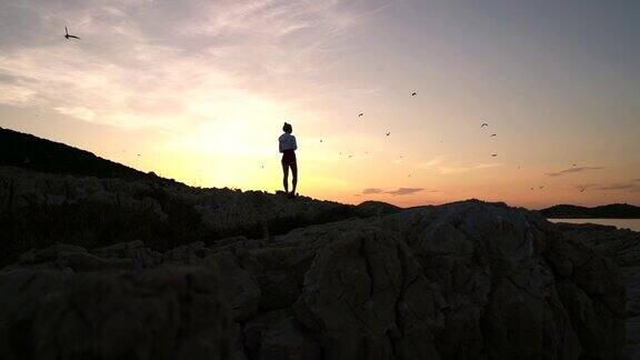 年轻女子在日落时站在岸上的岩石上看海鸥的慢动作