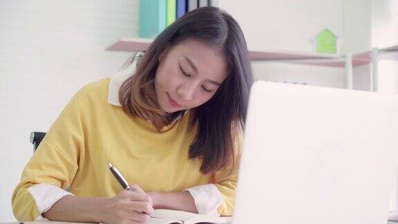 美丽的年轻微笑的亚洲妇女工作笔记本电脑在客厅在家里亚洲商务女性写笔记本文件财务和计算器在家庭办公室享受居家时光