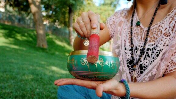 夏天在公园里一个女人坐在粉红色的瑜伽垫上玩歌唱碗古董视美丽的女孩拿着麻辣珠在绿草上沉思慢动作