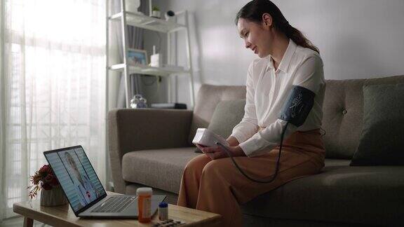 一位亚洲妇女病人在家里用笔记本电脑和她的医生聊天同时使用血压检查
