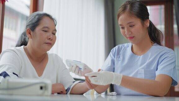 年轻的亚洲女性护工、护士、医生戴着防护手套坐在桌子上以积极的情绪检查亚洲老年糖尿病妇女的血糖