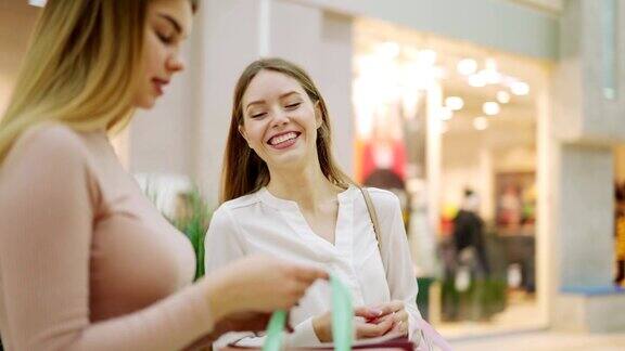 两个快乐的年轻女人在购物袋里互相展示购买的东西站在购物中心谈笑风生用平移放大镜头