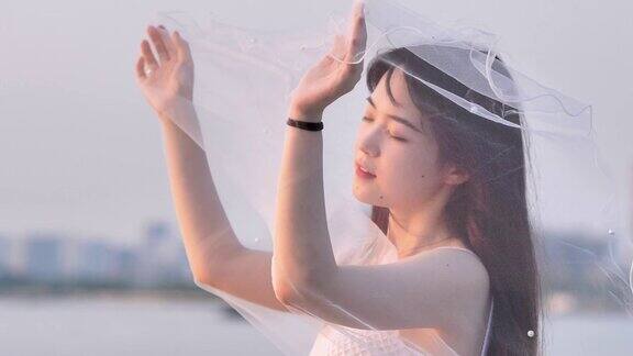 美丽的中国女孩在白色婚纱摆姿势闭上眼睛和面纱在风的黄昏与模糊的河水背景美丽浪漫4k镜头慢动作