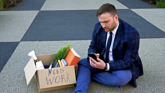 伤心的中年经理在丢掉工作后带着他的私人物品坐在地上人们试图通过互联网找工作