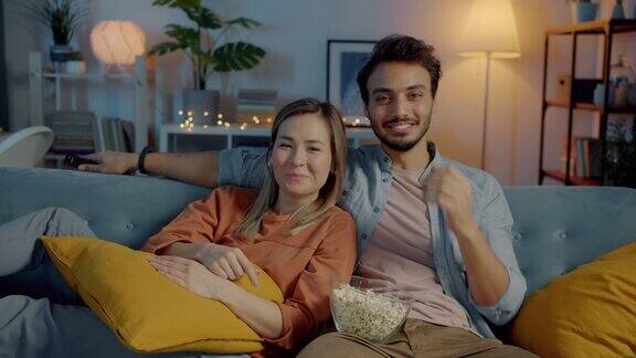 男朋友和女朋友的肖像看有趣的电影在电视上吃爆米花和笑在晚上在家