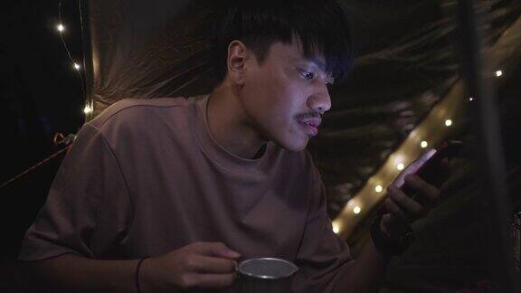 一个年轻人在晚上露营一边喝咖啡一边用他的智能手机