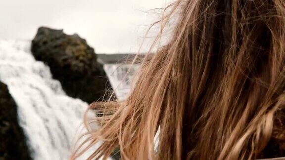 年轻美丽悲伤的女人站在冰岛的瀑布附近环顾四周的肖像头发随风摆动