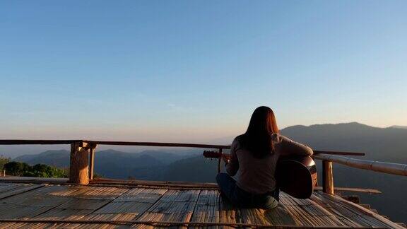 后视图的一个女性坐在和弹吉他在木阳台上与美丽的山景日落前
