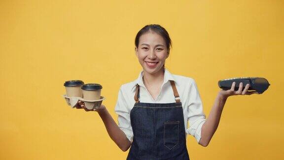 快乐微笑的亚洲女服务员穿着围裙友好地看着相机给顾客咖啡或茶和信用卡读卡机孤立在黄色背景