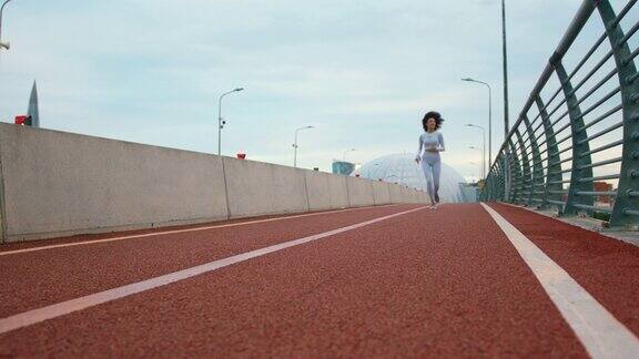 一个年轻健康的女运动员穿着白色紧身裤和黑色卷发正在桥上慢跑