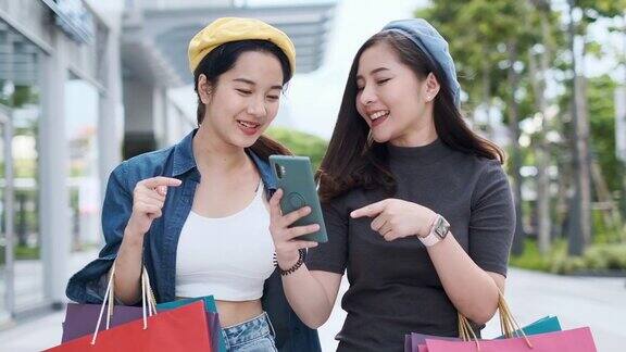 微笑年轻的亚洲女人与购物彩袋在商场背景用智能手机在网上购物站在商场大楼里微笑生活方式慢镜头4k