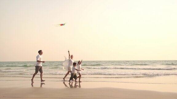 一个微笑的亚洲家庭的父母和两个孩子的男孩和女孩在夏天日落的海滩上一起跑和玩风筝的慢镜头快乐的家庭放松享受假期旅行的乐趣