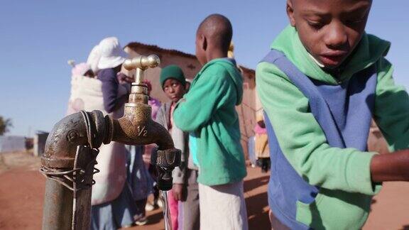 在南非由于严重的干旱年轻的非洲男孩从水龙头里喝水而妇女在排队收集塑料容器里的水