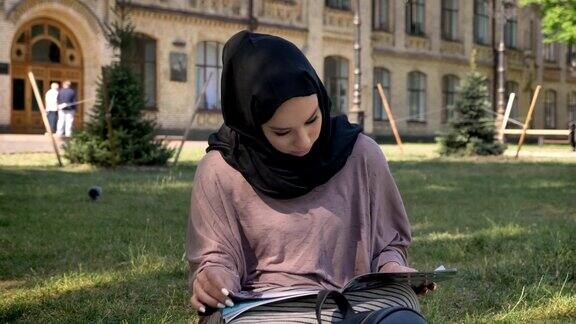 年轻的穆斯林女孩戴着头巾坐在草坪上看杂志建筑背景、宗教观念、放松观念