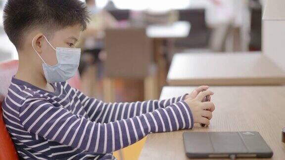 亚洲小男孩在图书馆学习在线手机戴口罩预防冠状病毒covid-19概念技术教育