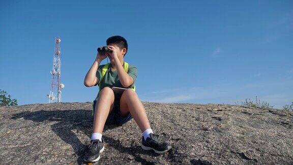 快乐的亚洲背包男孩在早上坐在山顶用望远镜看风景家庭徒步