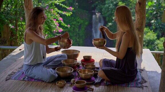 一名精通亚洲圣药的女子为一位年轻女性客户表演藏式碗治疗仪式的超慢镜头用西藏唱碗冥想他们在一个凉亭里冥想背景是美丽的瀑布