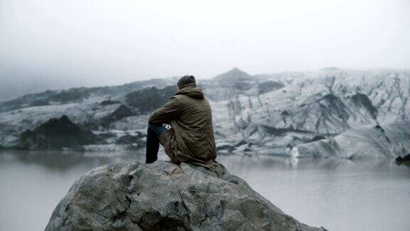 年轻男子独自坐在火山岩上看着冰岛的冰泻湖冰川的背影