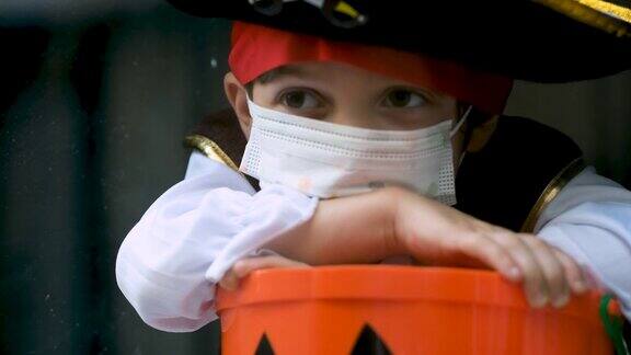 一个悲伤忧郁的小男孩穿着万圣节的海盗服装戴着保护面罩拿着一个杰克灯桶从窗户往外看