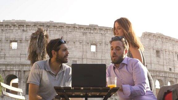 夕阳西下时三个年轻人坐在罗马圆形大剧场前的酒吧餐厅餐桌前用笔记本电脑在外面一起工作进行视频通话